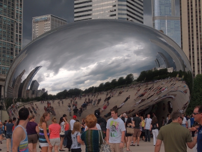[Large, silver, metal sculpture shaped like a bean. The surface reflects all around it including the sky, buildings, and many people viewing it.]